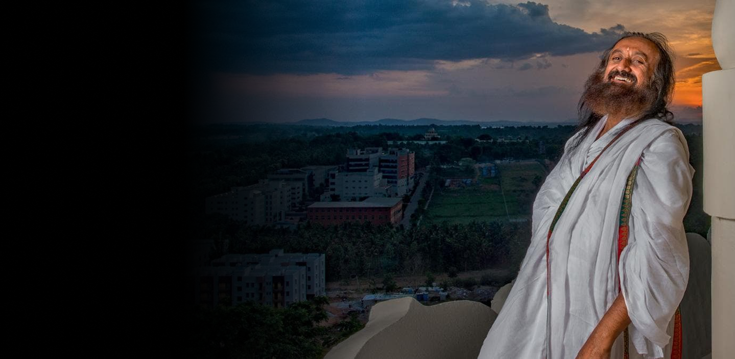 Sri Sri Ravi Shankar, founder of Art of Living, smiling in white robes against a dramatic sunset backdrop overlooking a campus with buildings and greenery