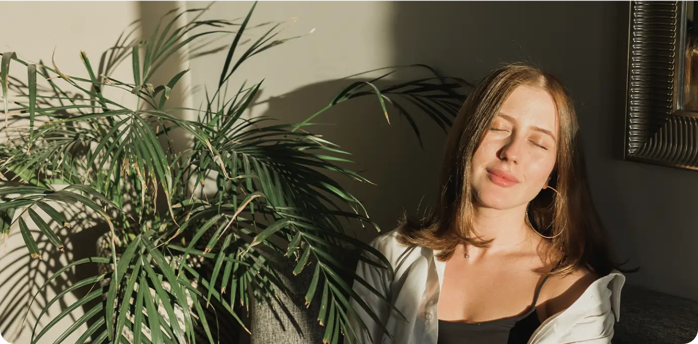 A woman sits on a couch, eyes closed, holding a mug, bathed in sunlight. Representing calm and relaxation from a deep sleep and anxiety relief course