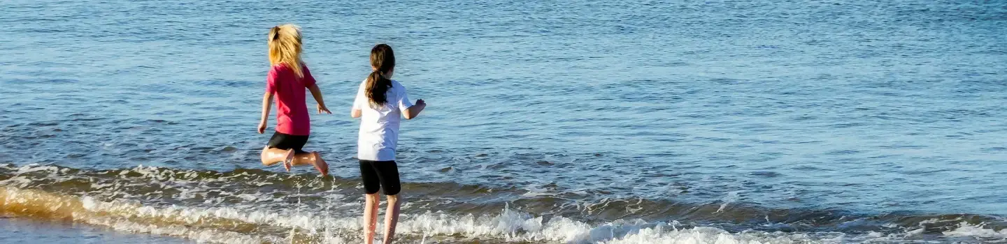 Duas crianças a brincar à beira-mar, chapinhando em ondas suaves, mostrando as competências de vida do ART Excel.