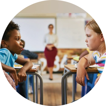 Two young girls smiling and chatting at a classroom desk, practicing communication skills.
