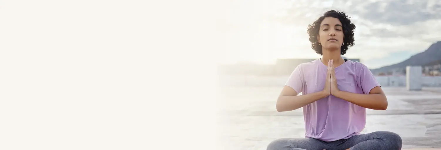 Une personne vêtue d'une chemise violette, assise les jambes croisées à l'extérieur, les yeux fermés et les mains en position de prière pour le programme Shakti Kriya.