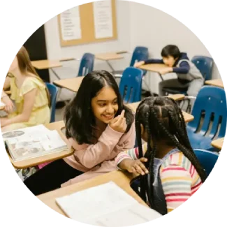 Two young girls smiling and chatting at a classroom desk, practicing communication skills.