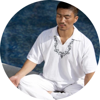A man dressed in white sits cross-legged near a body of water, with eyes closed and meditating