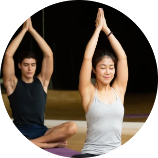 A woman and man sit cross-legged in a yoga studio, both with eyes closed and arms raised in a prayer position above their heads, practicing mindfulness and posture