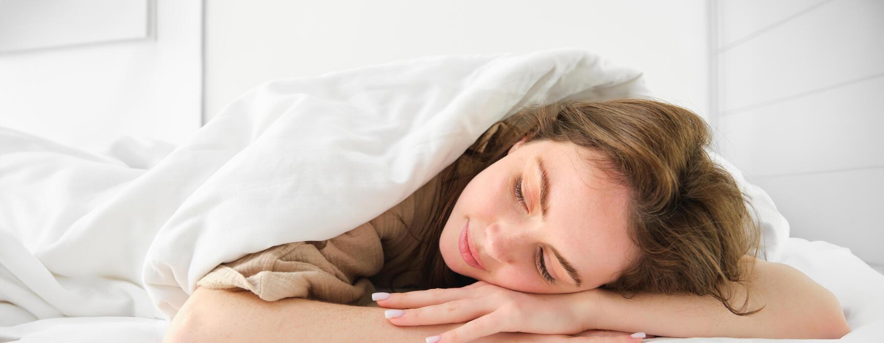 A woman sits on a couch, eyes closed, holding a mug, bathed in sunlight. Representing calm and relaxation from a deep sleep and anxiety relief course