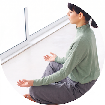 A woman dressed in white sits cross-legged with eyes closed and meditating in a bright room