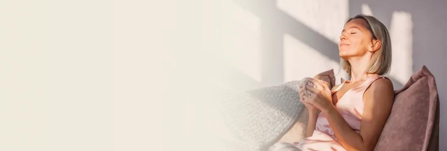 A woman sits on a couch, eyes closed, holding a mug, bathed in sunlight. Representing calm and relaxation from a deep sleep and anxiety relief course