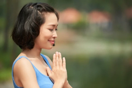 A serene woman in a blue top meditates with her hands in a namaste position, eyes closed and smiling slightly.
