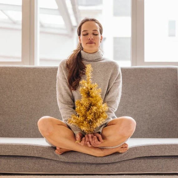 Woman meditating on a sofa, holding a small golden Christmas tree