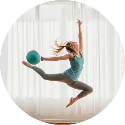 Žena skače s plavom loptom za vežbanje iznad glave, prikazujući dinamičnu, samouverenu pozu.