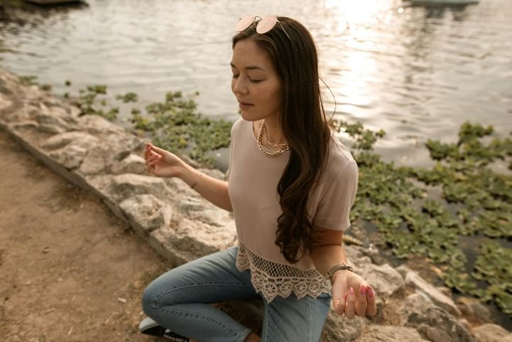 A young woman sitting outside, near water, practicing breathing exercises to relieve stress.