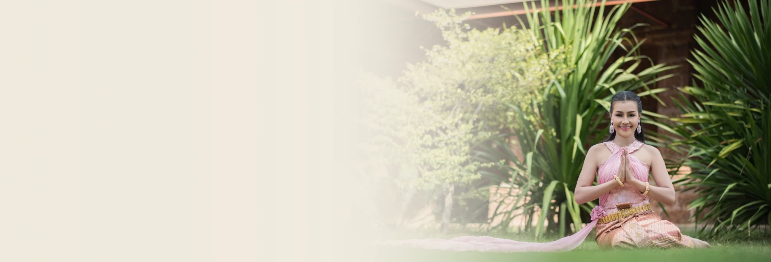 Một người phụ nữ mỉm cười trong bộ trang phục truyền thống màu hồng của Thái Lan đang ngồi trên bãi cỏ, thực hiện động tác chào 'wai'.