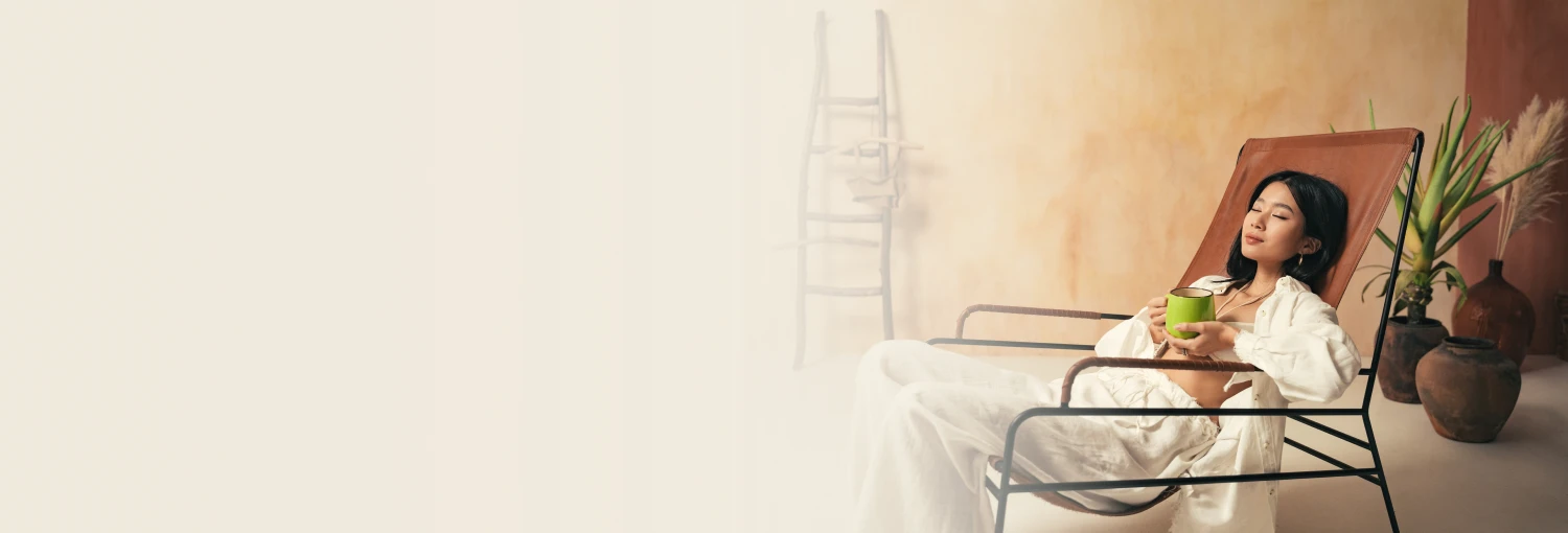 Một người phụ nữ ngồi trên ghế dài, nhắm mắt, tay cầm một chiếc cốc, tắm mình trong ánh nắng mặt trời. Hình ảnh này tượng trưng cho sự bình tĩnh và thư giãn sau một giấc ngủ sâu và liệu trình giảm lo âu.