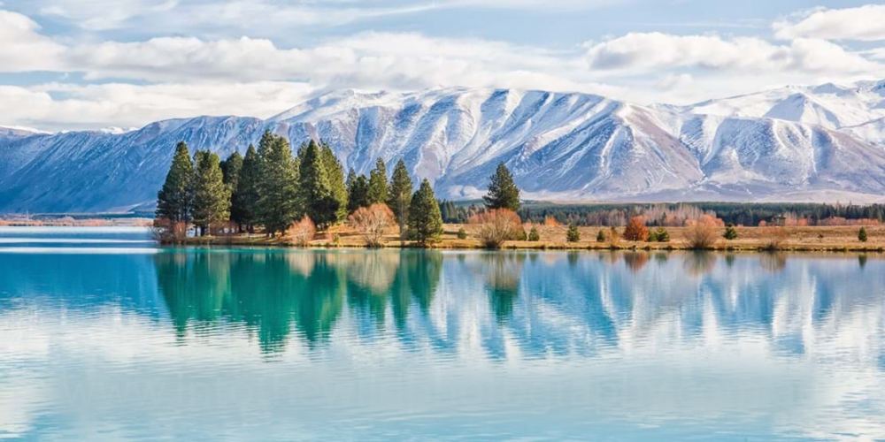 Serene landscape of a tranquil Lake Ruataniwha reflecting snow-capped mountains and lush trees under a partly cloudy sky.