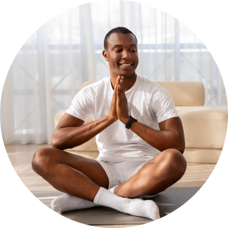 A man with open smile, sitting with legs crossed and hands in prayer position in a living area